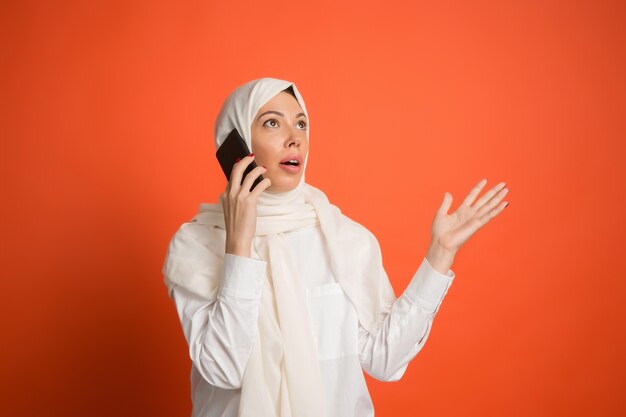 Feliz mujer árabe en hijab con teléfono móvil. Retrato de niña sonriente, posando en el fondo rojo del estudio.