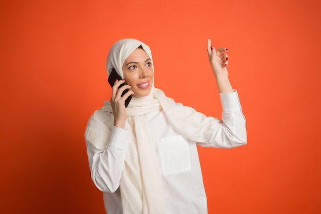 Feliz mujer árabe en hijab con teléfono móvil. Retrato de niña sonriente, posando en el fondo rojo del estudio.