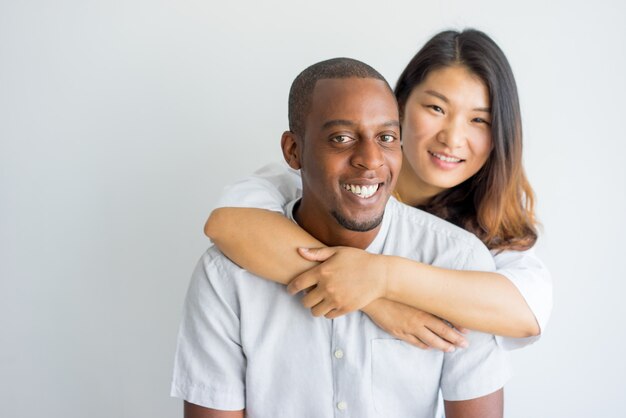 Feliz mujer asiática abrazando chico africano guapo y mirando a cámara.