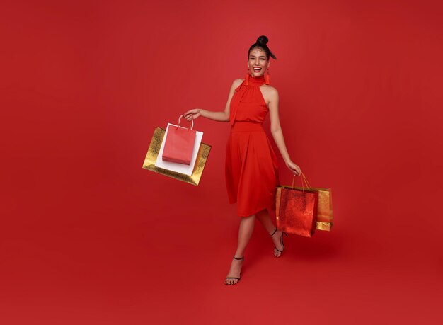 Feliz mujer asiática adicta a las compras con vestido rojo sosteniendo una bolsa de compras a pie del centro comercial