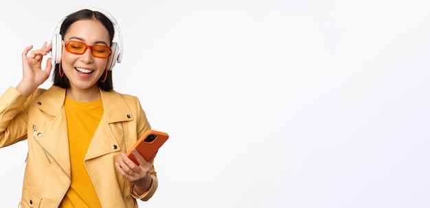 Feliz mujer asiática con estilo en gafas de sol escuchando música en auriculares con teléfono inteligente cantando y bailando de pie sobre fondo blanco.