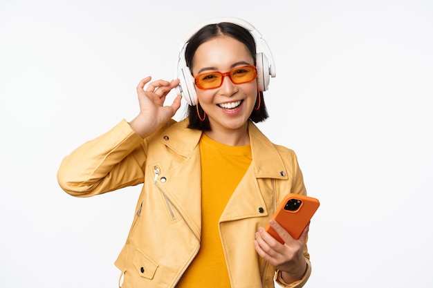 Feliz mujer asiática con estilo en gafas de sol escuchando música en auriculares con teléfono inteligente cantando y bailando de pie sobre fondo blanco.