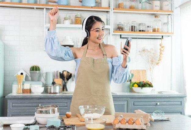 Feliz mujer asiática levanta el teléfono inteligente puño buen humor usar auriculares escuchar música en la cocina en casa