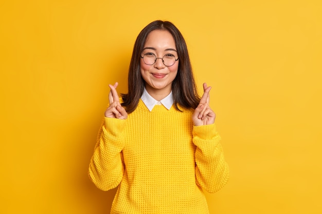 Feliz mujer asiática de pie con los dedos cruzados cree en la buena suerte en el examen espera que los sueños se hagan realidad usa gafas redondas y un suéter informal.