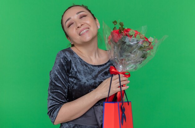 Feliz mujer asiática con ramo de rosas rojas y bolsa de papel con regalo sonriendo alegremente celebrando el día internacional de la mujer de pie sobre la pared verde