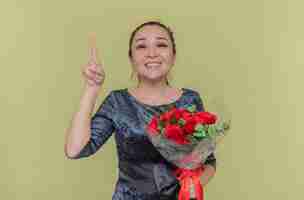 Foto gratuita feliz mujer asiática con ramo de rosas rojas sonriendo alegremente mostrando el dedo índice teniendo una nueva idea celebrando el día internacional de la mujer de pie sobre la pared verde