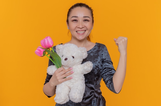 Feliz mujer asiática con ramo de tulipanes rosas y oso de peluche sonriendo alegremente apretando el puño celebrando el día internacional de la mujer de pie sobre una pared naranja