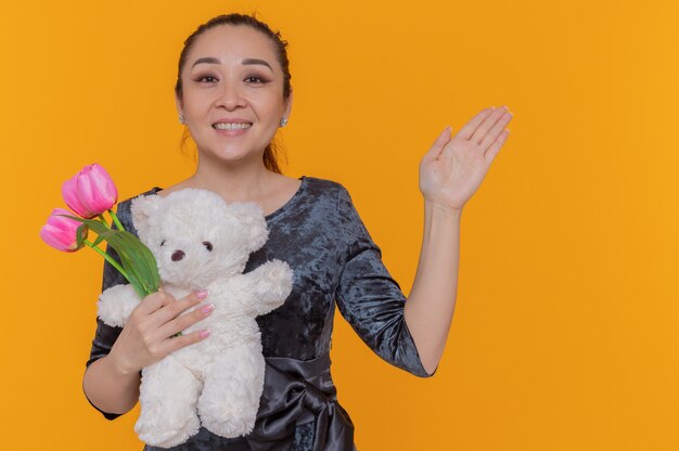 Feliz mujer asiática con ramo de tulipanes rosas y oso de peluche sonriendo alegremente celebrando el día internacional de la mujer de pie sobre la pared naranja