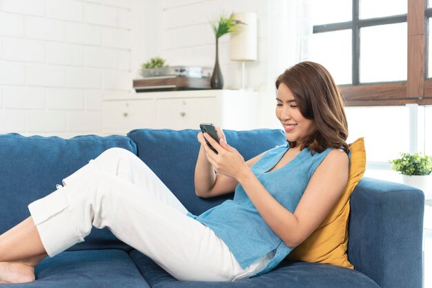 Feliz mujer asiática relajándose en un cómodo sofá con smartphone charlando en las redes sociales, viendo videos divertidos en casa.