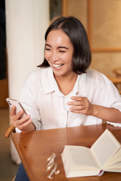 Feliz mujer asiática se sienta en un café con una taza de café y responde una videollamada en un teléfono inteligente riéndose