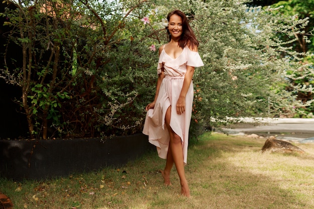 Feliz mujer asiática con sincera sonrisa en vestido rosa posando en el jardín de verano