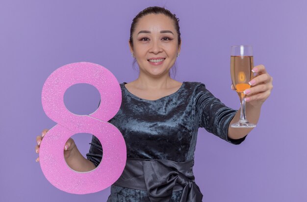 Feliz mujer asiática sosteniendo el número ocho de cartón y copa de champán sonriendo alegremente celebrando el día internacional de la mujer de pie sobre la pared púrpura