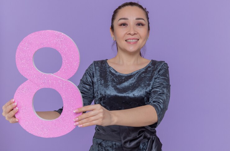 Foto gratuito feliz mujer asiática sosteniendo el número ocho de cartón mirando al frente sonriendo alegremente celebrando el día internacional de la mujer de pie sobre la pared púrpura