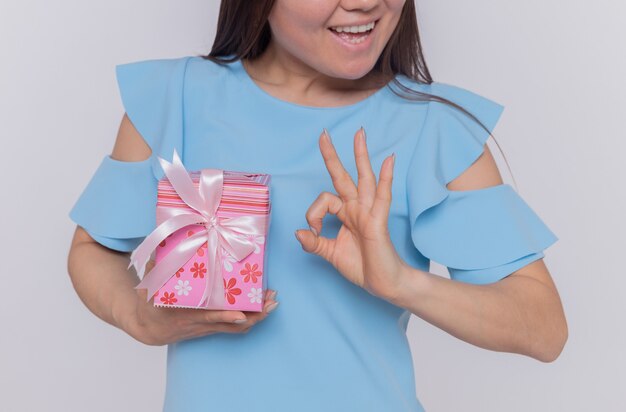 Feliz mujer asiática en vestido azul sosteniendo presente mostrando signo ok sonriendo celebrando el día internacional de la mujer de pie sobre una pared blanca