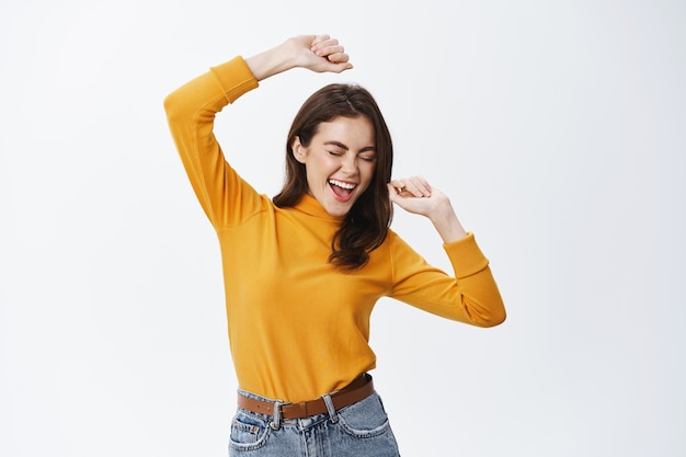 Feliz mujer atractiva bailando y divirtiéndose, levantando las manos sin preocupaciones, disfrutando de la música, de pie contra la pared blanca