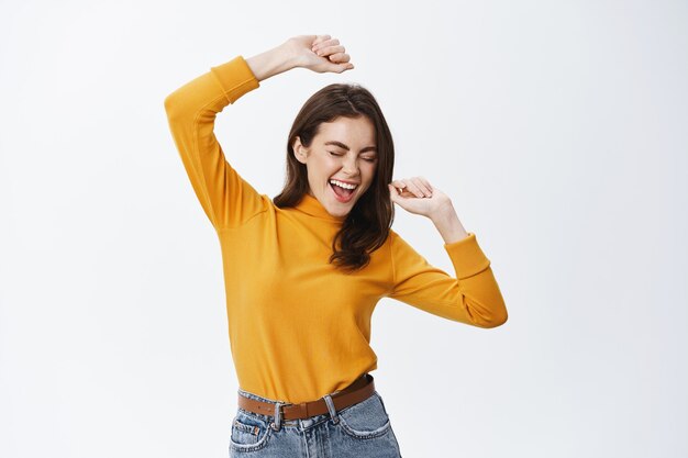 Feliz mujer atractiva bailando y divirtiéndose, levantando las manos sin preocupaciones, disfrutando de la música, de pie contra la pared blanca