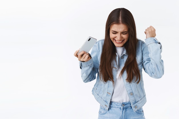 Feliz mujer atractiva logrando el objetivo de bombear el puño y celebrando la victoria mientras juega un juego móvil levantando las manos y saltando de la felicidad sosteniendo el teléfono inteligente horizontalmente Concepto de tecnología