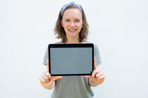 Feliz mujer bastante joven que muestra la pantalla en blanco de la tableta