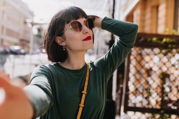 Feliz mujer blanca con pelo corto haciendo selfie en buen día de primavera. Foto al aire libre de chica interesada en elegantes gafas de sol y suéter verde.