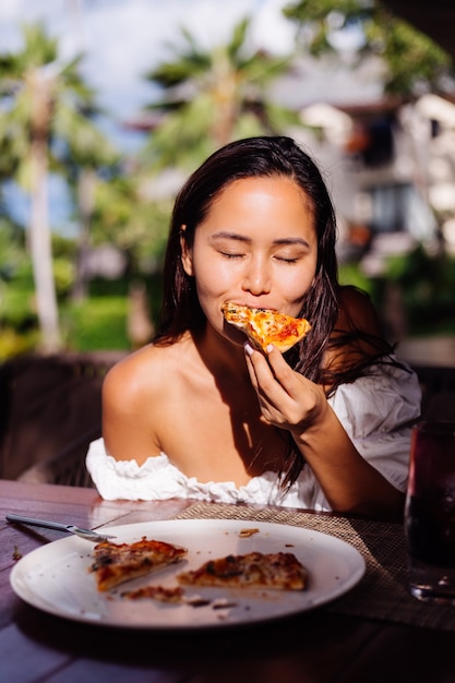 Foto gratuita feliz mujer bonita asiática hambrienta con pizza en el día soleado luz del atardecer en el restaurante al aire libre mujer disfrutando de la comida divirtiéndose en el almuerzo