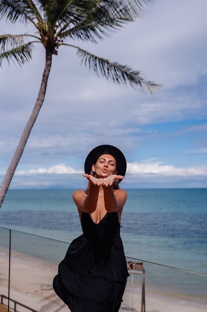 Feliz mujer bonita caucásica de vacaciones sonriendo en vestido negro y sombrero clásico, palmera y mar