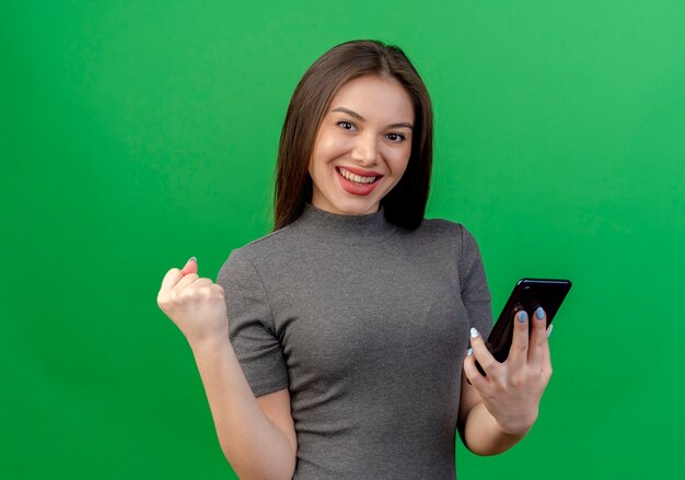 Feliz mujer bonita joven sosteniendo el teléfono móvil y apretando el puño aislado sobre fondo verde