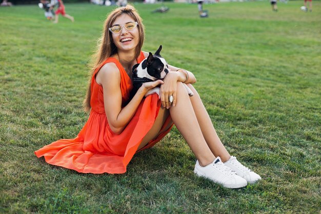 Feliz mujer bonita sentada en el césped en el parque de verano, sosteniendo el perro boston terrier, sonriendo con humor positivo, vestido naranja, estilo moderno, piernas delgadas, zapatillas de deporte, jugando con la mascota