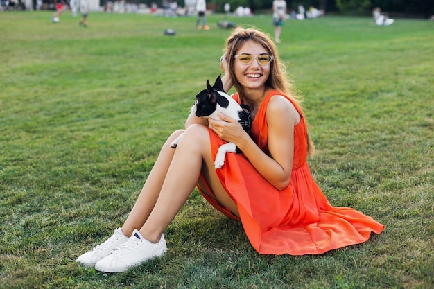 Feliz mujer bonita sentada en el césped en el parque de verano, sosteniendo el perro boston terrier, sonriendo con humor positivo, vestido naranja, estilo moderno, piernas delgadas, zapatillas de deporte, jugando con la mascota