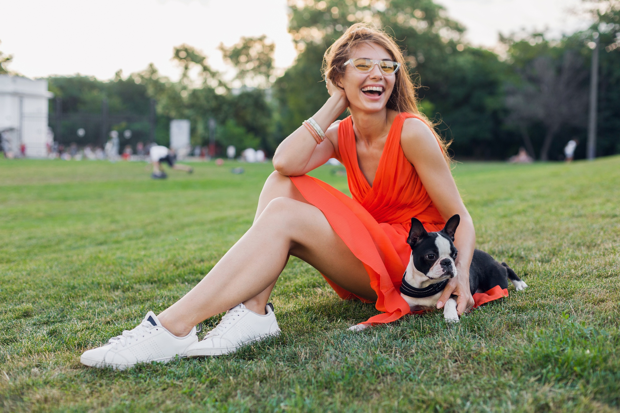 Feliz mujer bonita sentada en el césped en el parque de verano, sosteniendo el perro boston terrier, sonriendo con humor positivo, vestido naranja, estilo moderno, piernas delgadas, zapatillas de deporte, jugando con la mascota