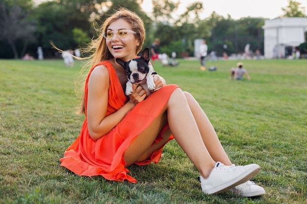 Feliz mujer bonita sentada en la hierba en el parque de verano, sosteniendo un perro boston terrier, sonriendo con humor positivo, vestido naranja, estilo moderno, piernas delgadas, zapatillas de deporte, jugando con la mascota, entretenimiento de fin de semana