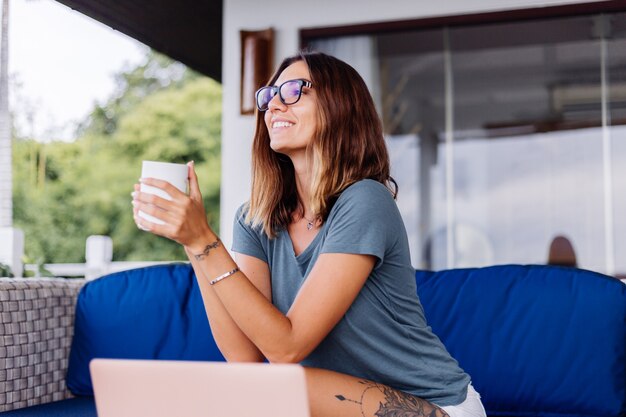 Feliz mujer caucásica hace trabajo a distancia en el portátil en casa lugar cómodo bebiendo té