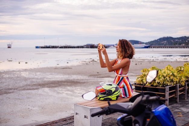 Feliz mujer caucásica del motorista en colorido vestido de verano de vacaciones con casco de moto