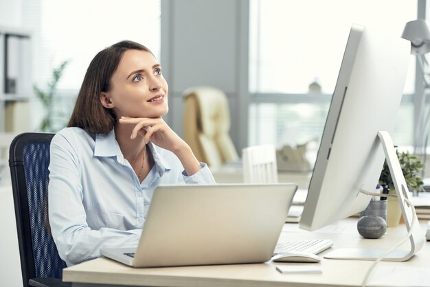 Feliz mujer caucásica soñando en la oficina frente a la computadora portátil y pantalla grande