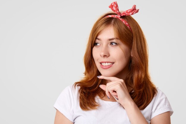 Feliz mujer caucásica con sonrisa positiva, sostiene la barbilla y mira alegremente a un lado, lleva una elegante diadema