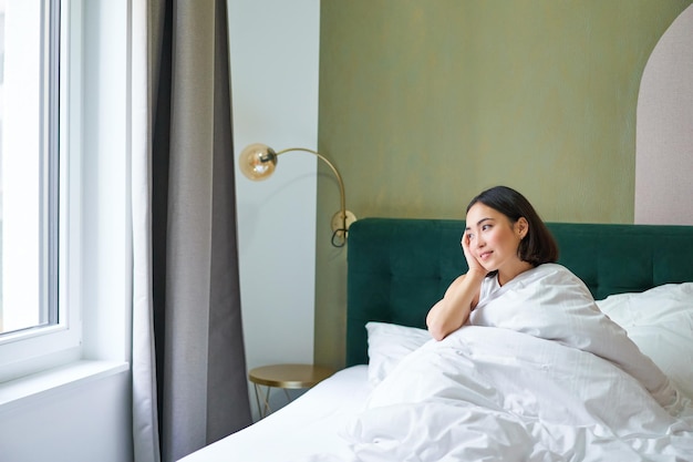 Foto gratuita feliz mujer coreana se despierta en su habitación de hotel acostada en la cama bajo acogedoras mantas cálidas sábanas blancas enj