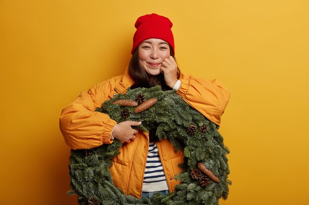 Feliz mujer coreana vestida con ropa de abrigo de invierno, expresa emociones sinceras, sostiene una hermosa corona de abeto, contra el fondo amarillo interior.