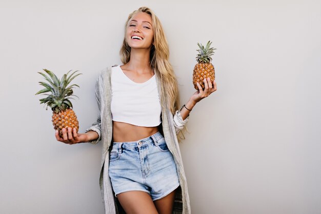 Feliz mujer curtida sosteniendo piñas y riendo. mujer con frutas exóticas aisladas sobre fondo claro.