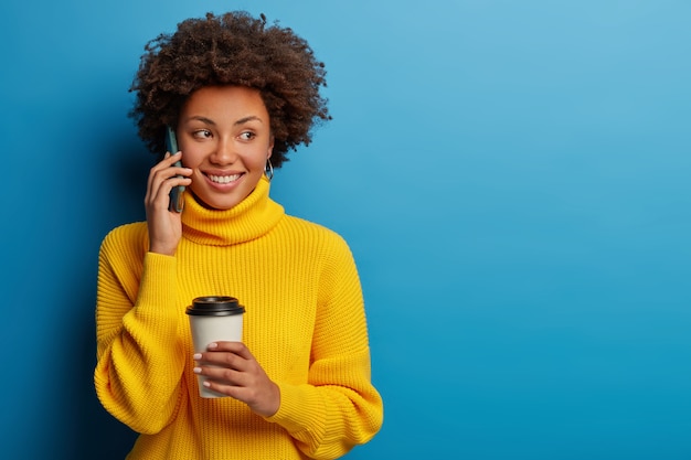 Feliz mujer despreocupada habla a través del teléfono móvil mientras bebe café, viste un suéter amarillo