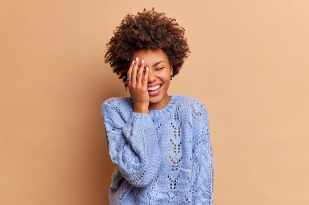 Feliz mujer despreocupada con el pelo rizado se ríe a carcajadas de una broma divertida hace que la palma de la cara vea algo hilarante viste un jersey casual aislado sobre una pared beige