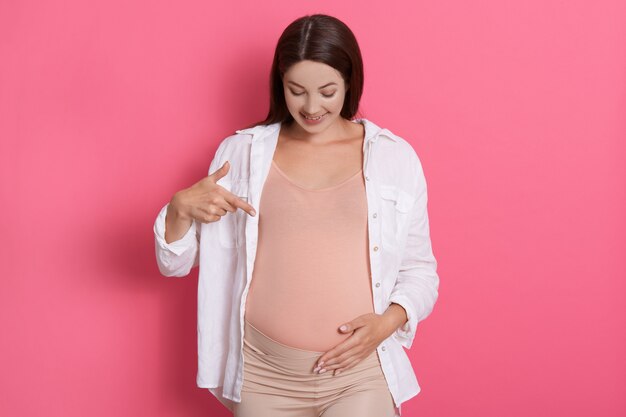 Feliz mujer embarazada apuntando a su vientre con una sonrisa y expresión facial positiva, vestida con camisa blanca mientras está de pie contra la pared rosa, madre esperada de cabello oscuro.