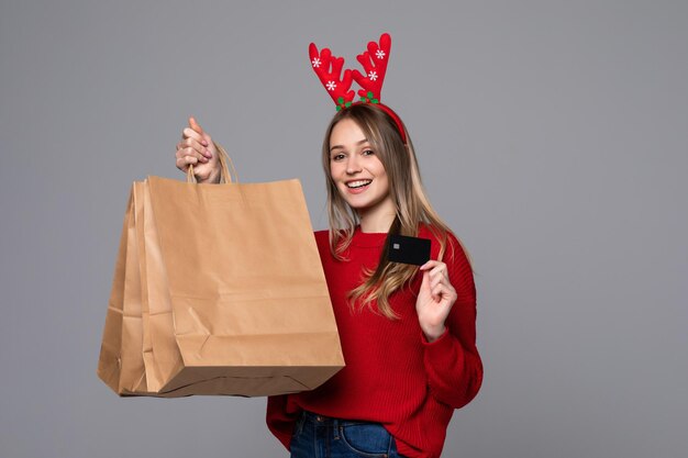 Foto gratuita feliz mujer encantadora con cuernos de reno en su cabeza bolsas de compras con tarjeta de crédito en la pared gris