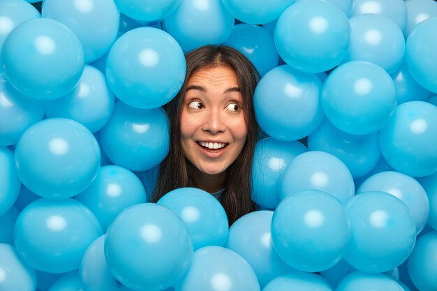 Feliz mujer étnica mira misteriosamente a un lado y una amplia sonrisa posa contra globos azules esperando un evento especial.