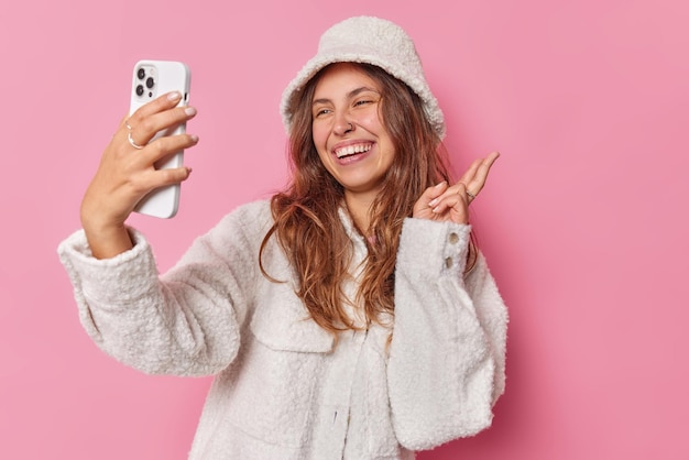 Feliz mujer europea de cabello largo hace un gesto de paz y se toma selfie en el teléfono móvil sonríe ampliamente vestida con ropa de invierno de moda aislada sobre fondo rosa disfruta de una videollamada con un amigo