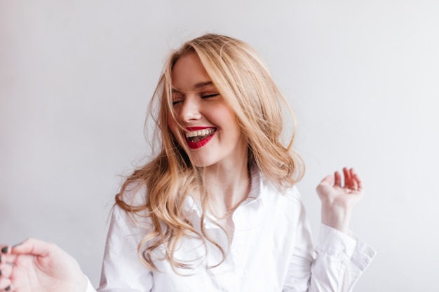 Feliz mujer europea con camisa blanca expresando emociones positivas. alegre chica rubia en la pared de luz.