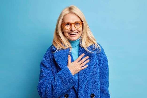 Feliz mujer europea rubia senior divertida con broma humorística ríe positivamente mantiene la mano en el pecho vestida con abrigo azul de invierno.