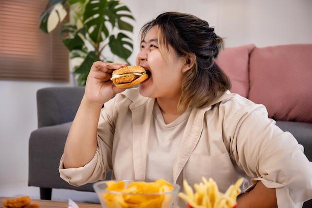 Feliz mujer gorda asiática disfruta comiendo deliciosas hamburguesas en la sala de estar