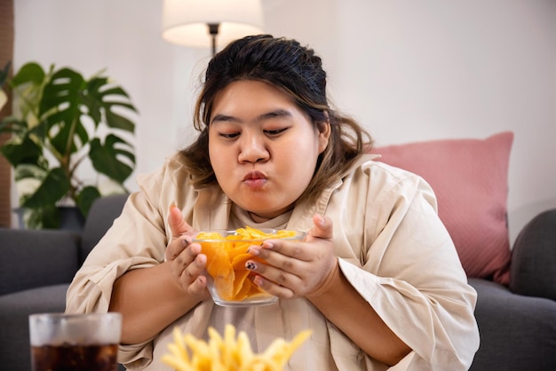 Feliz mujer gorda asiática disfruta comiendo deliciosas papas fritas y papas fritas en la sala de estar