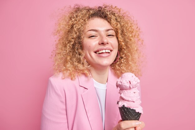 Feliz mujer guapa con cabello rizado disfruta comiendo sabroso helado de fresa tiene una expresión alegre estilo de vida despreocupado expresa emociones auténticas vestidas elegantemente aisladas sobre una pared rosa