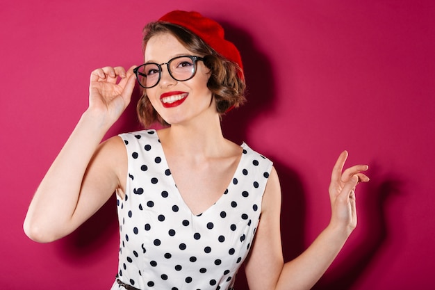 Feliz mujer de jengibre en vestido y anteojos mirando a la cámara sobre rosa