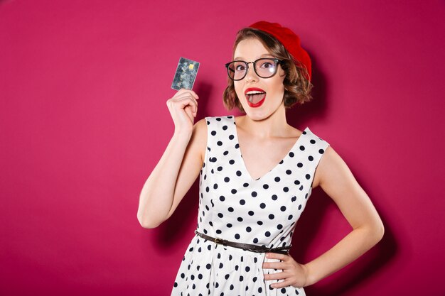 Feliz mujer de jengibre en vestido y anteojos con tarjeta de crédito mientras mira a la cámara sobre rosa